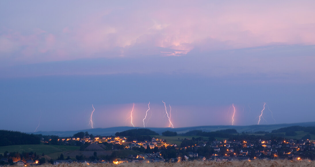 Fernblick auf eine Gemeinde, im Hintergrund dunkle Wolken und viele Blitze, die in die Erde einschlagen